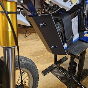 Könnte beinhalten: Eine schwarze Batteriebox aus Metall mit einem schwarzen Riemen und einem weißen Etikett mit Text. Die Batteriebox ist an einem blau-weißen Dirtbike befestigt.