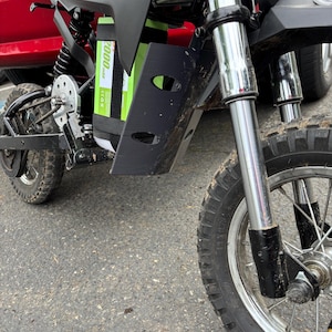 May include: Black and silver electric mini-bike with a green battery pack. The bike has a black seat, a front headlight, and a black mudguard. The tires are black with a knobby tread. The bike is parked on asphalt.