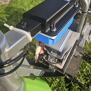 Puede incluir: Un conjunto de componentes de vehículos eléctricos con una carcasa rectangular negra asegurada con pernos. Un componente azul y blanco es visible debajo de la carcasa negra. El conjunto está montado en un marco plateado, con piezas verdes y negras también visibles.