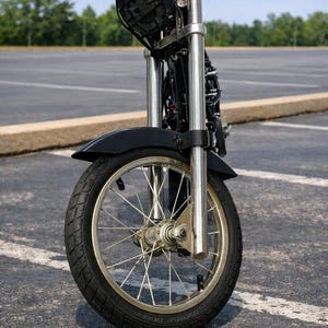 Op de afbeelding: Vooraanzicht van een kleine, op maat gemaakte motorfiets. De motorfiets heeft een zwarte band, zilveren spaken en een zwart spatbord. De voorvorken zijn zilver en de koplamp is zwart. De motorfiets staat op asfalt.