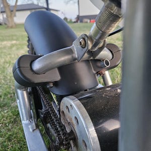May include: A close-up view of a small electric motor, featuring a black body and silver chain and sprocket. The motor is mounted on a silver frame with a black fender. Green grass and a house are in the background.