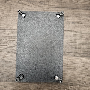 Puede incluir: Una placa rectangular de metal negro texturizado con cuatro pernos en cada esquina. Los pernos son plateados y negros. La placa está sobre una superficie de madera.