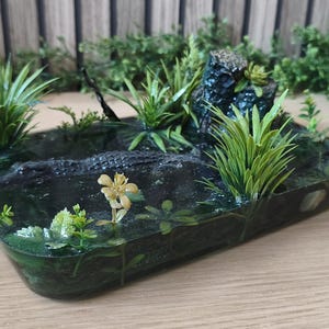 Może przedstawiać: Prostokątna diorama z żywicy przedstawiająca mroczną scenę bagienną z zatopioną figurką aligatora, różnymi zielonymi sztucznymi roślinami wodnymi, małą formacją skalną i pojedynczym żółtym kwiatem, zatopionymi w przezroczystej, ciemnozielonej żywicy.