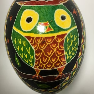 May include: A hand-painted Easter egg with a black background and a colorful owl design. The owl has green feathers, yellow eyes, and a red beak. The egg is decorated with a geometric pattern in yellow, green, and red.