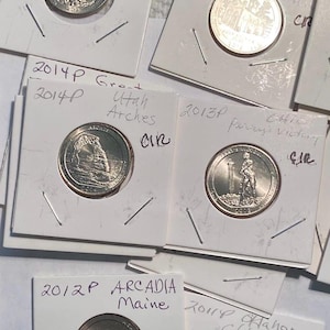 May include: A collection of U.S. quarter coins, each encased in a protective holder. The coins feature various designs and are labeled with the state and year of minting, including Massachusetts, Utah, and Maine. The collection is a numismatic item.