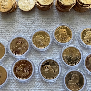 May include: A collection of gold-colored Sacagawea dollar coins, some loose and some encased in clear plastic protective capsules. The coins feature the profile of Sacagawea, an eagle in flight, and agricultural imagery with the text Liberty and 2000.