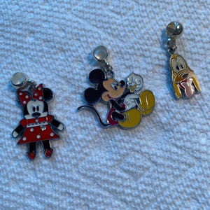 May include: Three Disney-themed charms featuring beloved characters. Minnie Mouse is in a red dress with white polka dots. Mickey Mouse is in red shorts and yellow shoes. Pluto has a yellow body and pink tongue.