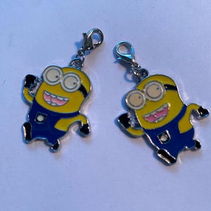 May include: Two Minion-themed charms. Each charm features a yellow body, blue overalls, and black shoes. The charms have silver-colored clasps for attachment. The Minions have large eyes and are smiling with pink mouths.
