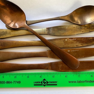 May include: A collection of vintage copper-toned flatware, including spoons and knives. The knives have serrated edges and the word "Signature" is etched on the blades. A green ruler with white text is at the bottom.
