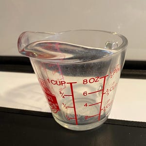 May include: A clear glass measuring cup with red markings for ounces and cups. The cup has a curved handle and a pouring spout. The measurements are clearly visible, with a maximum capacity of 1 cup or 8 ounces.