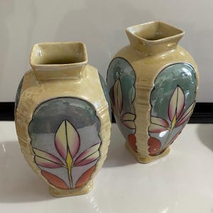 May include: Two decorative vases with a square top and a rounded body. The vases are beige with a floral design in pink, green, and orange. The vases have a glossy finish and are likely made of ceramic.