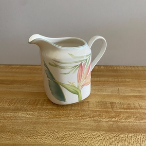 May include: White ceramic creamer with a handle and spout, featuring a floral design with pink, green, and orange tropical flowers and leaves. The rim has a light blue trim. The creamer is likely for serving coffee or tea.