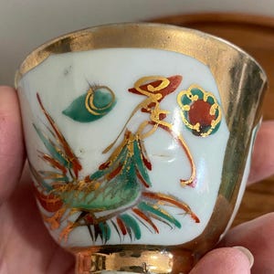 May include: Small, white ceramic cup with a gold rim and base. The cup is decorated with a colorful design featuring a bird with green, red, and gold accents. The design is hand-painted, and the cup appears to be vintage.