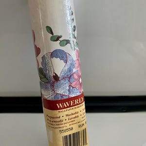 May include: Rolled wallpaper border with a floral design in pink, blue, and green. The label reads "Waverly Wallpaper Borders" and includes the words "Prepasted" and "Washable". The roll is wrapped in clear plastic and has a blue stripe at the top and bottom.