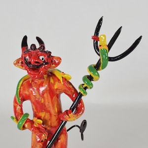 May include: A red devil figurine with horns and a tail, holding a black trident with a green snake wrapped around it. The devil is painted with orange and yellow accents.