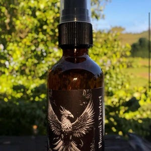 Puede incluir: Una botella de vidrio marrón con un atomizador negro y una etiqueta en blanco y negro con un diseño de fénix. La etiqueta dice "Phoenix Protect".