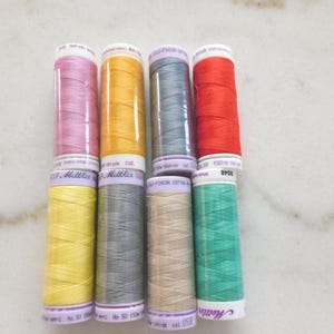 May include: Eight spools of Mettler sewing thread arranged in two rows of four. The colors include pink, yellow, grey, red, light yellow, dark grey, beige, and teal. Each spool features white labels with black text indicating 150 meters of thread.