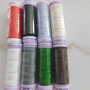 May include: Eight spools of Mettler Silk-Finish cotton sewing thread arranged in two rows. The colors include red, black, light blue, cream, white, grey, green, and dark brown. Each spool features a light purple label with printed text.