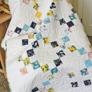 May include: A white quilt with a patchwork design featuring squares of various floral and geometric patterns in shades of blue, yellow, pink, and black. The quilt is quilted with a swirling pattern.
