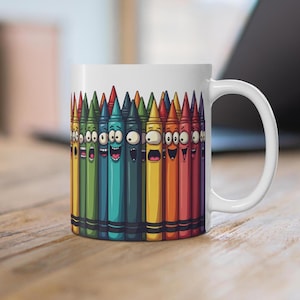 May include: White ceramic mug featuring a colourful design of cartoon crayons with faces. The crayons are arranged in a row and are all different colours.