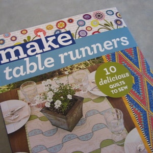 May include: Book cover with the title "Make table runners" and a colorful floral design. The book features 10 quilt patterns to sew.