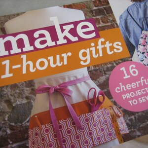 May include: A book cover with the text "make 1-hour gifts" in white letters on a pink and orange background. The cover also features a pink circle with the text "16 cheerful projects to sew". An apron and scissors are visible.