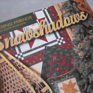 May include: A book cover titled "Snowshadows" by Kathryn Squibb & Deborah Jacobs. The cover features a quilt design with red, green, and white patterns. A snowshoe and evergreen branches are also visible, suggesting a winter theme.