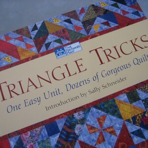 May include: Book cover with a colorful patchwork quilt design. The title is "Triangle Tricks: One Easy Unit, Dozens of Gorgeous Quilts" with an introduction by Sally Schneider.
