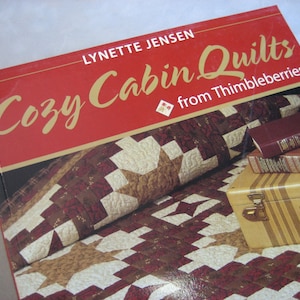 May include: A book titled "Cozy Cabin Quilts from Thimbleberries" by Lynette Jensen. The cover features a red, white, and brown quilt with a star pattern.
