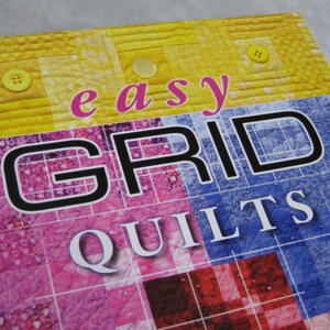 May include: A book cover with the title "easy GRID QUILTS" in black and white text on a colorful patchwork background. The background features squares of yellow, pink, red, and blue fabric.