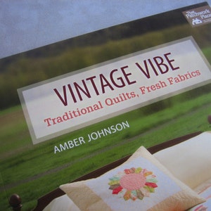 May include: A book cover with a green field and a bed with pillows. The title is "Vintage Vibe Traditional Quilts, Fresh Fabrics" by Amber Johnson. The book cover is from The Patchwork Place.