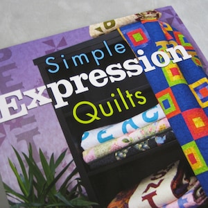 May include: Book cover with the title "Simple Expression Quilts" in white text on a black background. The cover features a bookshelf with colorful quilts stacked on the shelves. The background is a purple and white pattern.