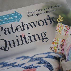 May include: A book titled "Visual Guide to Patchwork & Quilting" with the subtitle "Fabric Selection to Finishing Techniques & Beyond". The cover features a blue arrow with white polka dots and the words "Visual Guide To" on it. The book cover also features a stack of colorful fabric scraps and a basket with more fabric scraps.