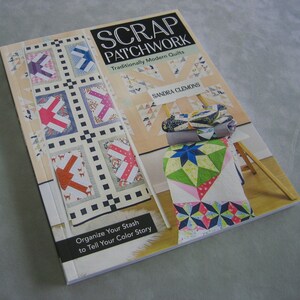May include: Book cover with the title "Scrap Patchwork: Traditionally Modern Quilts" by Sandra Clemons. The cover features a quilt with a patchwork design in various colors and patterns. The book also includes a photo of a quilt on a wooden easel.