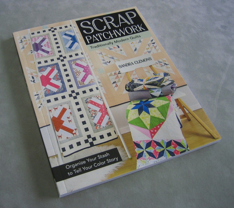 May include: Book cover with the title "Scrap Patchwork: Traditionally Modern Quilts" by Sandra Clemons. The cover features a quilt with a patchwork design in various colors and patterns. The book also includes a photo of a quilt on a wooden easel.