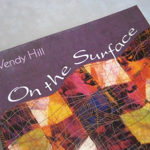 May include: Book cover with a purple background and the title "On the Surface" in white cursive text. The author's name, "Wendy Hill", is printed in white text above the title. The cover features a colorful patchwork design with a variety of textures.