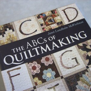 May include: The ABCs of Quiltmaking book cover features a dark blue background with white text. The title is "The ABCs of Quiltmaking" with the author's name "Janet Lundholm McWorkman" below. The cover features a patchwork quilt design with various colors and patterns.