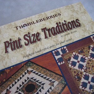 May include: Book cover for "Pint Size Traditions" by Lynette Jensen. The cover features a beige background with a floral pattern and the title in red and black lettering. The book is about making handcrafted quilts for small spaces.
