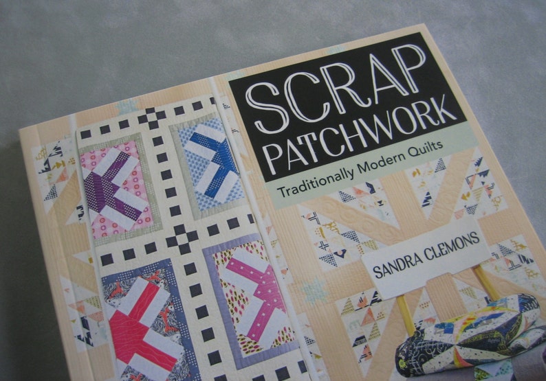 May include: A book titled "Scrap Patchwork: Traditionally Modern Quilts" by Sandra Clemons. The cover features a patchwork quilt design with a variety of colors and patterns. The book is a guide to making modern quilts using scrap fabric.