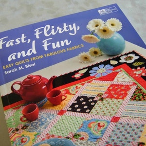 May include: A book titled "Fast, flirty, and fun: Easy quilts from fabulous fabrics" by Sarah M. Bisel. The book cover features a colorful patchwork quilt with a red teapot and teacups on a white surface. The background is a blue sky with white flowers.