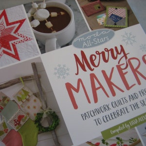 May include: A book cover with a red and white star, a ladder with stockings, and a mug of hot chocolate with marshmallows. The title of the book is "Merry Makers: Patchwork Quilts and Projects to Celebrate the Season" by Lissa Alexander.
