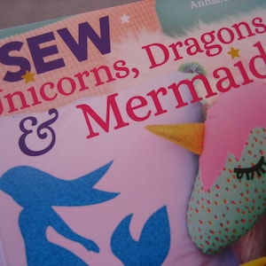 May include: A book cover with the title "Sew Unicorns, Dragons & Mermaids" in pink, purple, and yellow text. The cover features a pink and green unicorn with a yellow horn and a blue mermaid silhouette.