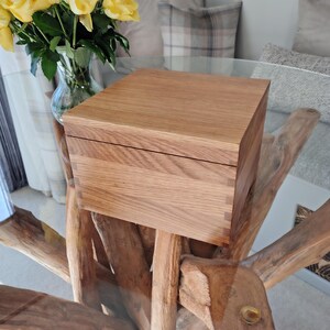 May include: A square wooden box with a natural light brown grain and visible finger joints at the corners. The box features a flat, smooth lid that sits flush with the sides, showcasing the texture and patterns of the wood grain throughout the exterior.