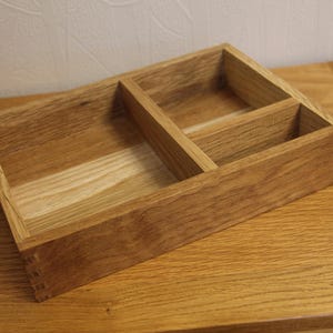 May include: A wooden storage box with three sections. The box is crafted from light brown wood and rests on a similar wooden surface. Internal dividers create three distinct compartments. The wood grain is clearly visible throughout the box's construction.
