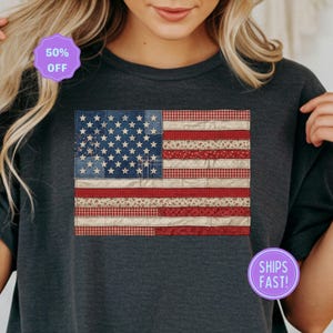 May include: Dark gray t-shirt featuring a patchwork American flag design. The flag incorporates denim, floral, and gingham patterns in its construction. The shirt has a round neckline and short sleeves. A purple sticker that says "50% OFF" is visible.