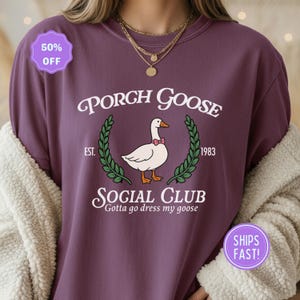 May include: A maroon sweatshirt featuring the text "PORCH GOOSE SOCIAL CLUB" and a goose graphic with a bow tie. The phrase "Gotta go dress my goose" is below the graphic. The sweatshirt also has the text "EST. 1983".