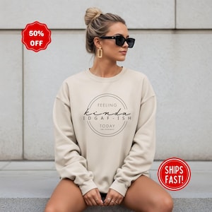 Puede incluir: Sudadera beige con el texto "Feeling Kinda IDGAF-ish Today" en un diseño circular negro. La sudadera tiene mangas largas y cuello redondo. Un círculo rojo con "50% OFF" está en la esquina superior izquierda.