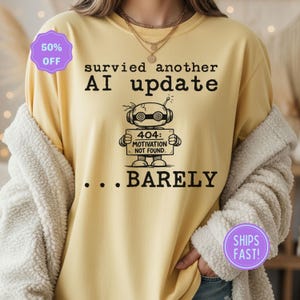 Puede incluir: Una camiseta de manga larga de color amarillo claro con el texto "survied another AI update" y un gráfico de robot que sostiene un letrero que dice "404: MOTIVATION NOT FOUND... BARELY". Una pegatina morada dice "50% OFF".