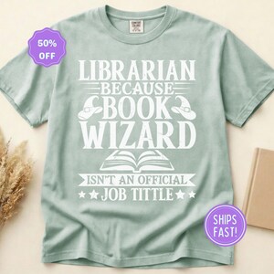 May include: A sage green short-sleeve t-shirt featuring white text that reads LIBRARIAN BECAUSE BOOK WIZARD ISN'T AN OFFICIAL JOB TITTLE, accompanied by illustrations of two wizard hats and an open book centered on the front of the garment.