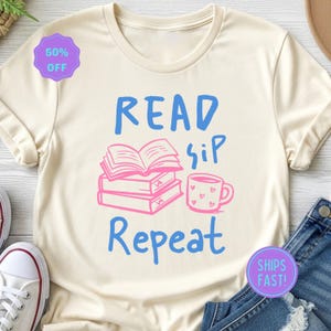 以下が含まれることがあります： クリーム色のTシャツ。青とピンクで「READ sip Repeat」の文字がプリントされています。デザインには本の山、ハートのマグカップ、テキストが含まれています。紫色のステッカーは50％オフを示しています。